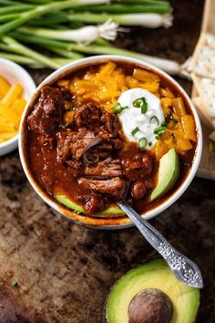 a bowl filled with meat and vegetables on top of a wooden table next to sliced avocado