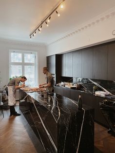 two men are preparing food at the counter in a large, white room with wood floors