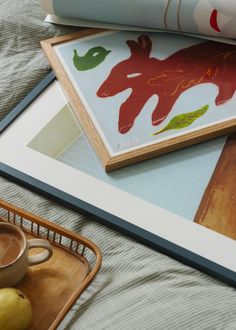 two framed pictures sitting on top of a bed next to an apple and coffee cup