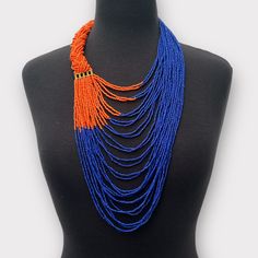 This beautiful beaded necklace features a long, orange and purple braided intricate beadwork. Its design reflects the traditional aesthetic of African fashion, with a touch of modern style. The hook closure secures the necklace, making it a convenient accessory for any occasion. The vibrant orange colour adds an eye-catching element to any outfit, while the beaded fringe adds a unique and elegant touch. This necklace is perfect for those who appreciate African jewellery's beauty and cultural sig Orange Multi-strand Beaded Necklaces, Orange Multi-strand Polished Beaded Necklaces, Orange Multi-strand Beaded Necklace, Orange Multi-strand Beaded Necklace With Polished Beads, Handmade Multi-strand Orange Beaded Necklaces, Orange Multi-strand Polished Beaded Necklace, Traditional Orange Faceted Beads, Orange Multi-strand Bohemian Beads, Bohemian Orange Faceted Bead Necklace