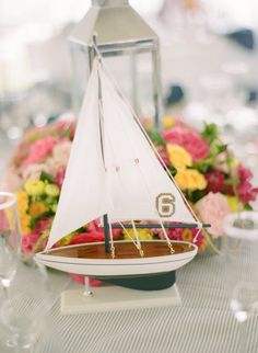 a model sailboat is sitting on a table with flowers and wine glasses in the background