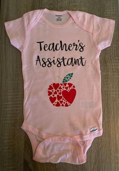 a pink baby bodysuit with an apple on the front and teacher's assistant written on it