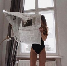 a woman in a black swimsuit reading a news paper while standing next to a window