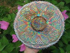 a hand holding a colorful glass plate in front of some purple and pink flowers with green leaves