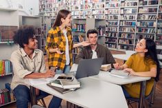 three people sitting at a table with laptops and books in a library, talking