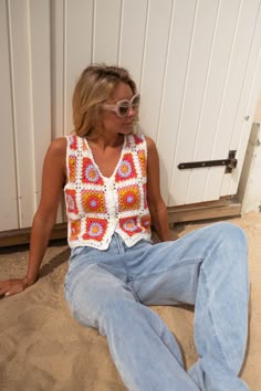 a woman sitting in the sand with her legs crossed wearing sunglasses and a crocheted top