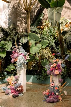 three white vases filled with colorful flowers on top of a stone floor next to palm trees