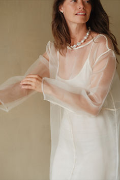 a woman wearing a white dress with sheer sleeves and pearls on her necklace, standing in front of a beige wall