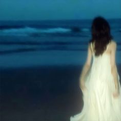 a woman in a white dress standing on the beach at night with her back to the camera