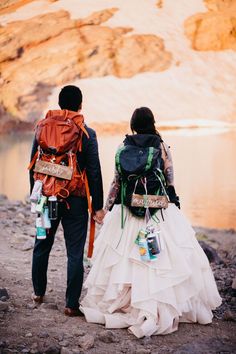 two people with backpacks walking down a path by the water in their wedding dresses