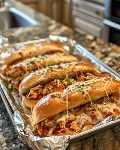 hot dogs in buns covered with cheese and toppings sitting on tin foil lined up on a kitchen counter