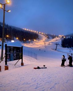 several people on skis at the bottom of a snowy slope with lights in the background