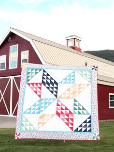 a quilt hanging from a pole in front of a barn with a red building and green grass