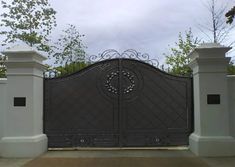 an iron gate in front of a house with trees and bushes behind it on a cloudy day