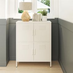 a white cabinet sitting in front of a window next to a lamp and a plant