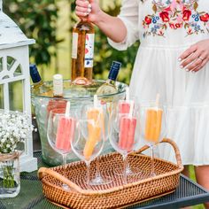 a woman holding a bottle and glasses filled with drinks on top of a picnic table