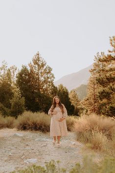 a woman in a dress standing on a dirt road surrounded by tall grass and trees