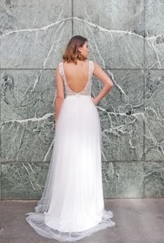 a woman standing in front of a wall wearing a white wedding dress with an open back