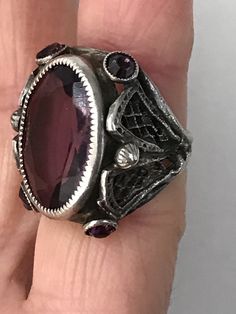 "This lovely filigreed and ornate ring has an oval shaped faceted amethyst purple color glass stone at its center. It is flanked on either side of to and bottom with small round bezel set purple glass stones. The sides have lattice shaped cut outs and the band is smooth. The face of this ring is about 3/4\" long and about 1/2\" across. It is a size 3 1/4 and is marked Sterling. Please note there are some overall signs of wear, especially to the stones, four of five of which, including the center Antique Oval Purple Jewelry, Antique Purple Oval Jewelry, Ornate Oval Amethyst Ring, Purple Oval Filigree Rings, Victorian Style Oval Purple Amethyst Ring, Silver Amethyst Ring With Intricate Design, Oval Amethyst Ring With Filigree Detail, Purple Oval Stone Jewelry, Purple Oval Stones Jewelry