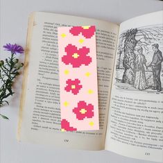 an open book with a pink and yellow pixel pattern on it next to a purple flower