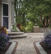 a brick walkway leading to a house with flowers in the foreground and trees on either side