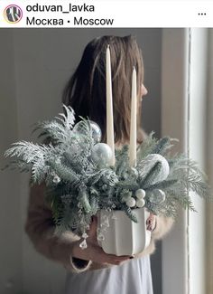 a woman holding a christmas arrangement with candles and greenery in her hands while standing next to a window