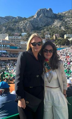 two women standing next to each other in front of a stadium full of people and mountains