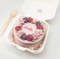 a pink cake with purple flowers on top in a plastic container next to a fork and spoon