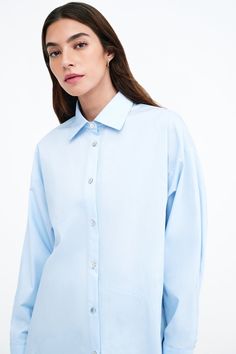 Our Ezra Shirt is a capsule wardrobe dream for your closet. Crafted from 100% crisp European poplin cotton, she offers a classic button-up silhouette in front, but with a cropped back for modern edge. Say hello, as well, to her diagonal seams at the front, sleek collar and long dolman sleeves finished with mother-of-pearl button cuffs. Wear Ezra tucked, tied or open and free—she never fails to inspire new looks.[SPLIT] Rocio, in light blue and in black, is 5'9.5" (175 cm) tall, wearing size XS. Chic Poplin Top With Relaxed Fit, Classic Relaxed Fit Button-up Cropped Shirt, Relaxed Fit Poplin Top For Work, Long Sleeve Poplin Tops With Button Closure, Oversized Poplin Tops For Spring, Relaxed Fit Poplin Tops With Button Cuffs, Summer Long Sleeve Poplin Shirt, Cotton Cropped Shirt With Button Cuffs, Cotton Cropped Shirt With Button Cuffs For Spring