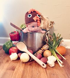 a baby sitting in a pot surrounded by fruits and vegetables, with a hat on