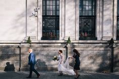 two people walking down the street in front of a building, one wearing a wedding dress