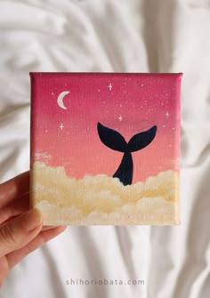 a hand holding up a small painting of a whale tail in front of a pink sky