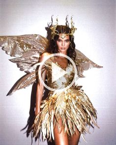 a woman in a gold and white costume with wings on her head, standing next to a wall