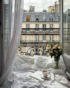 a vase with flowers sitting on top of a bed in front of a window next to a cup of coffee