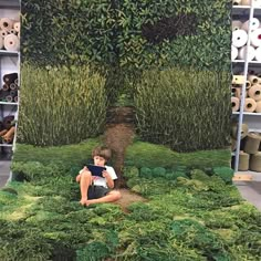 a woman sitting on the ground in front of a green wall with plants growing out of it