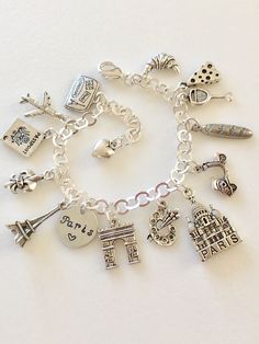 "Adorable Paris charm bracelet featuring 13 detailed silver charms of travel to Paris, classic landmarks, artist quarter, food and drink. Hand stamped 1/2\" 18 aluminum disc says \"Paris ❤️\". Memories of a visit abroad living \"la bonne vie\" in the magical city of Paris! Charms include: suitcase, plane, passport, fleur de lis, Eiffel Tower, Arc de Triumph, artist palette, Sacre Coeur church, Vespa scooter, baguette, cheese, wine, and croissant. Charms are antique silver, pewter and silver tone Silver Symbolic Jewelry Souvenir, Silver Charms Jewelry Souvenir, Silver Metal Jewelry For Travel, Nickel-free Silver Charms Souvenir, Nickel-free Silver Charms For Souvenir, Silver Engraved Jewelry For Travel, Silver Nickel-free Bracelet Souvenir, Nickel-free Silver Bracelets As Souvenir, Silver Charm Bracelet As Souvenir