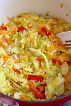 a red pot filled with cabbage and carrots on top of a table next to a wooden spoon