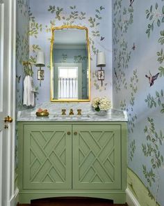 a bathroom with green cabinetry and wallpaper on the walls, along with a gold framed mirror