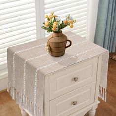 a vase with flowers on top of a white table next to a wooden drawer and window