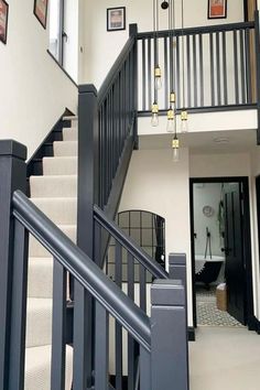 A modern staircase with Scandinavian-inspired design, featuring bold black railings and spindles paired with a soft, neutral herringbone runner. Hanging from the double-height ceiling are minimalist pendant lights with exposed bulbs and gold accents. The upper landing overlooks the hallway, where a large arched mirror leans against the wall. A glimpse into an adjacent bathroom reveals a classic black clawfoot tub and patterned floor tiles, enhancing the sleek and contemporary look.