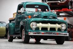 an old green truck parked in a garage next to other classic cars on the ground