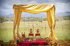 an outdoor wedding setup with red and yellow drapes, gold chairs and flowers on the table