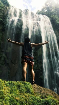 a person standing on top of a lush green hillside next to a waterfall with their arms outstretched