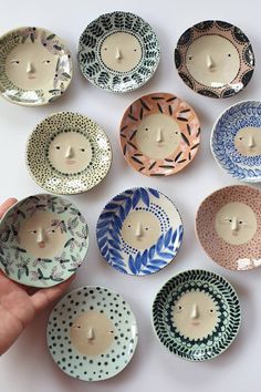 a hand is holding a small bowl with many designs on it and there are nine bowls in the shape of people's faces