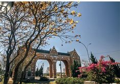 an arch in the middle of a park with trees and flowers around it on a sunny day