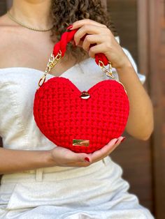 a woman holding a red crocheted heart shaped purse in her right hand and wearing a white dress