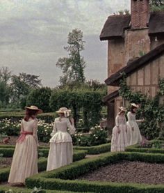 four women in dresses and straw hats are walking through a garden area with hedges, bushes and flowers