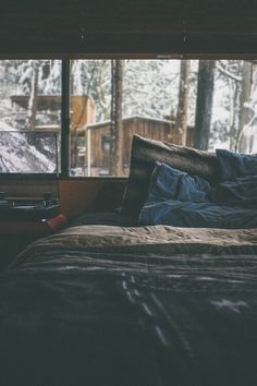 an unmade bed in front of a window with snow on the ground and trees outside