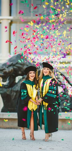 two women in graduation gowns standing next to each other with confetti falling around them