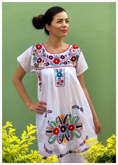 a woman standing in front of a green wall wearing a white dress with colorful flowers on it